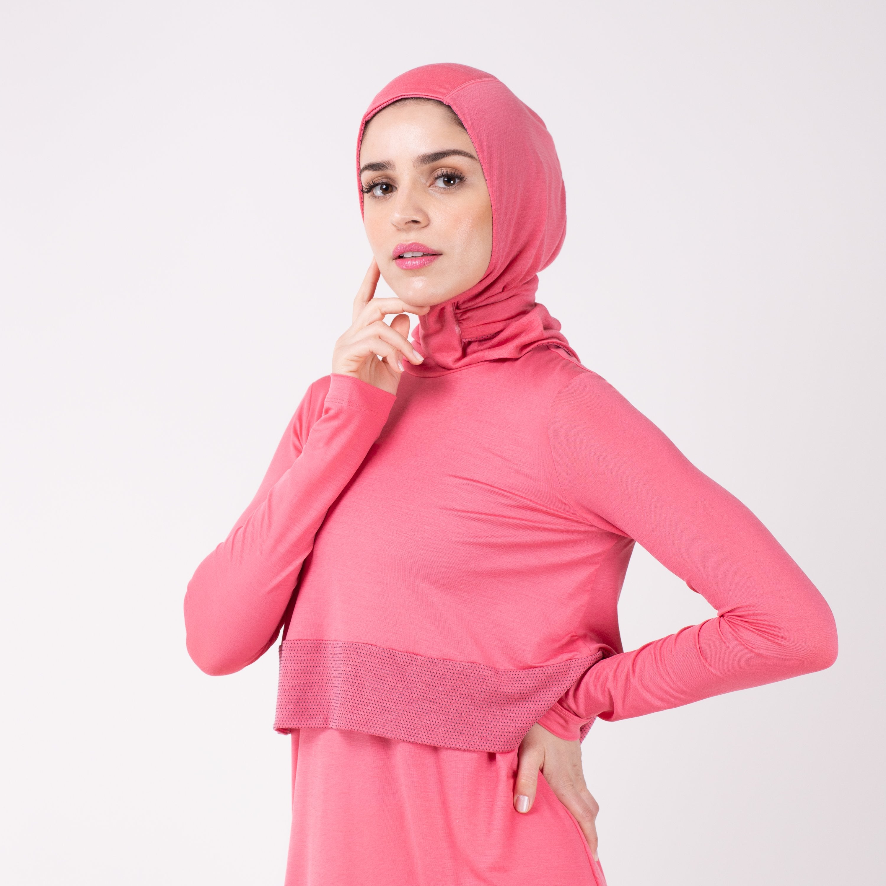 Woman facing left in a pink shirt with a matching pink HAWA hijab, touching her face with her right and and her left hand on her hip.