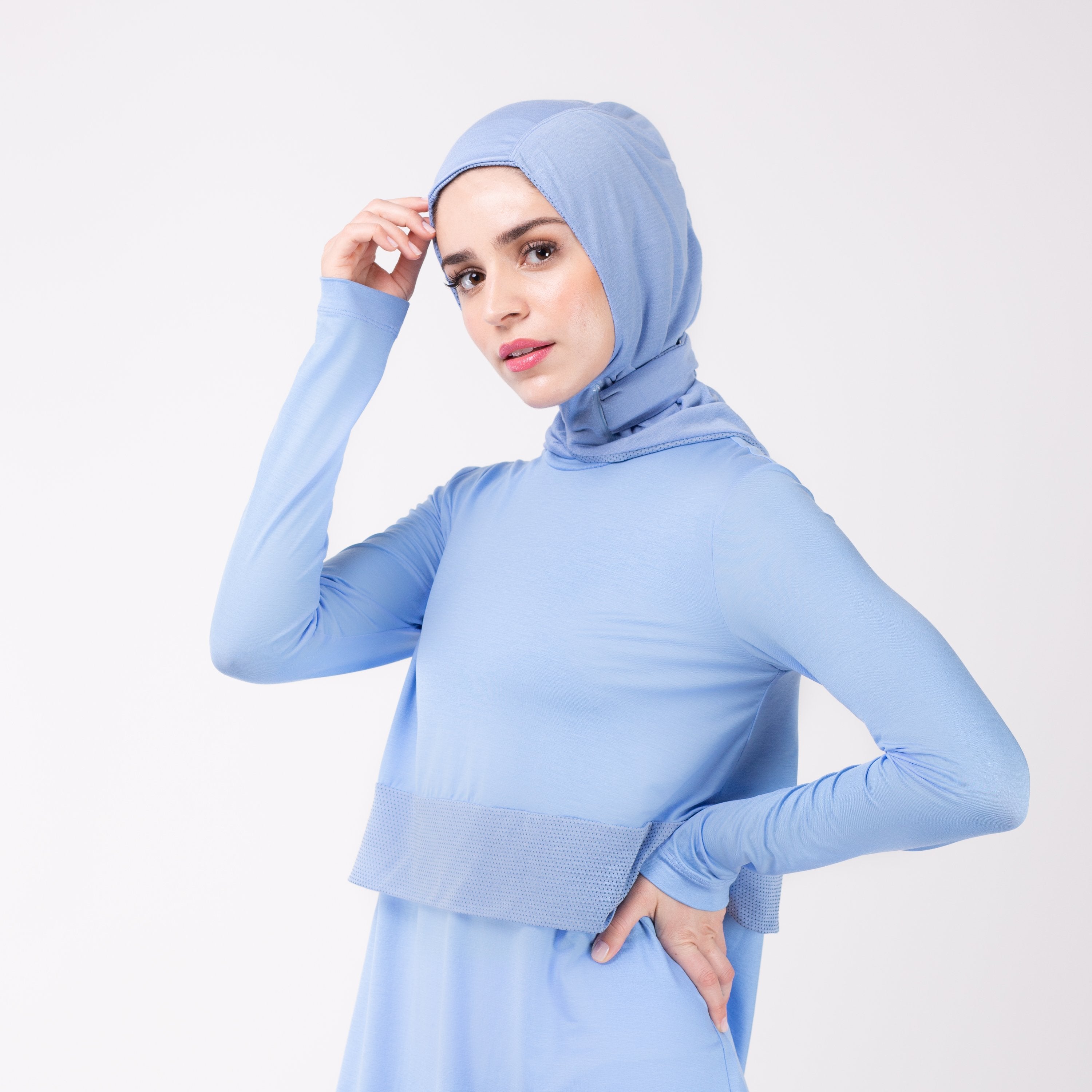 Woman facing left in a sky blue shirt with a matching sky blue HAWA hijab, touching the hijab fabric with her right and and her left hand on her hip.