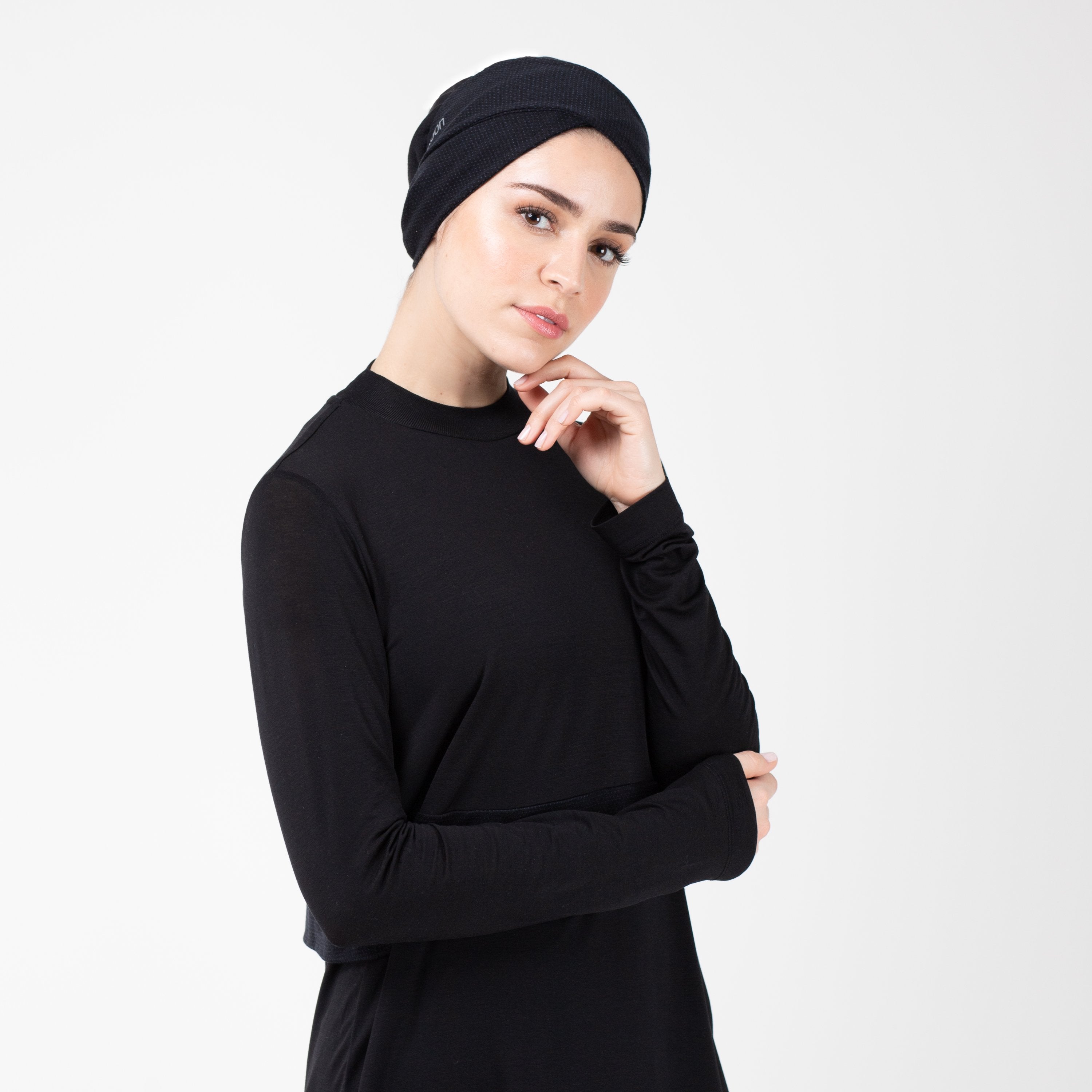 Woman facing right in a black shirt with matching black HAWA headwrap, touching her face with her left hand.