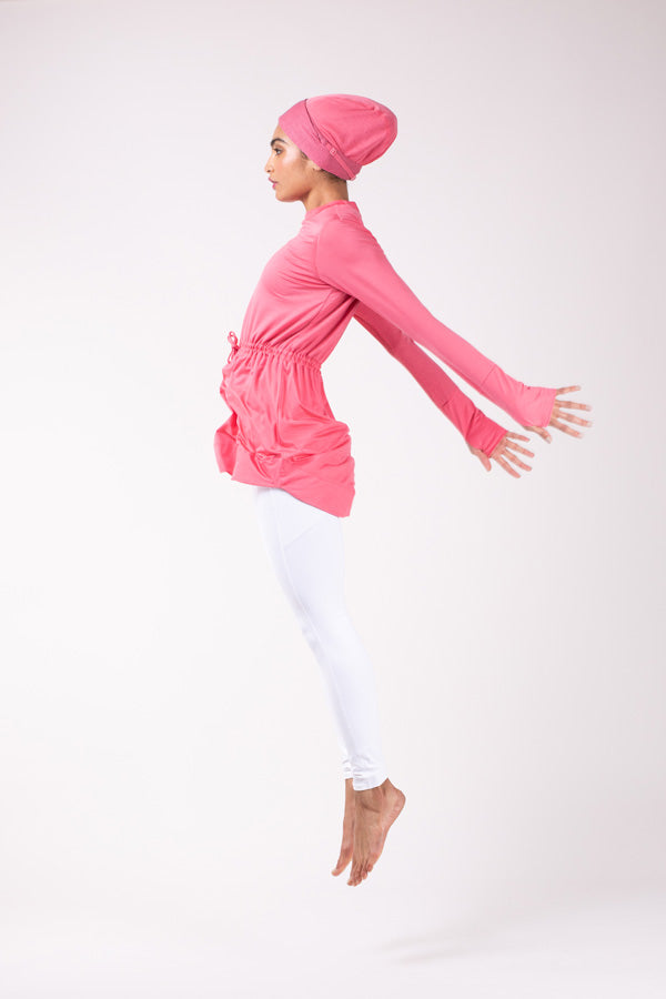 Woman jumping into the air in white pants, a pink HAWA drawstrip tee, and a pink HAWA headwrap.