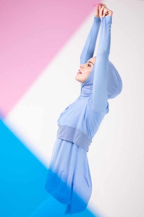 A woman leaning back with her arms in the air in a sky blue HAWA step tee with a matching sky blue HAWA hijab.