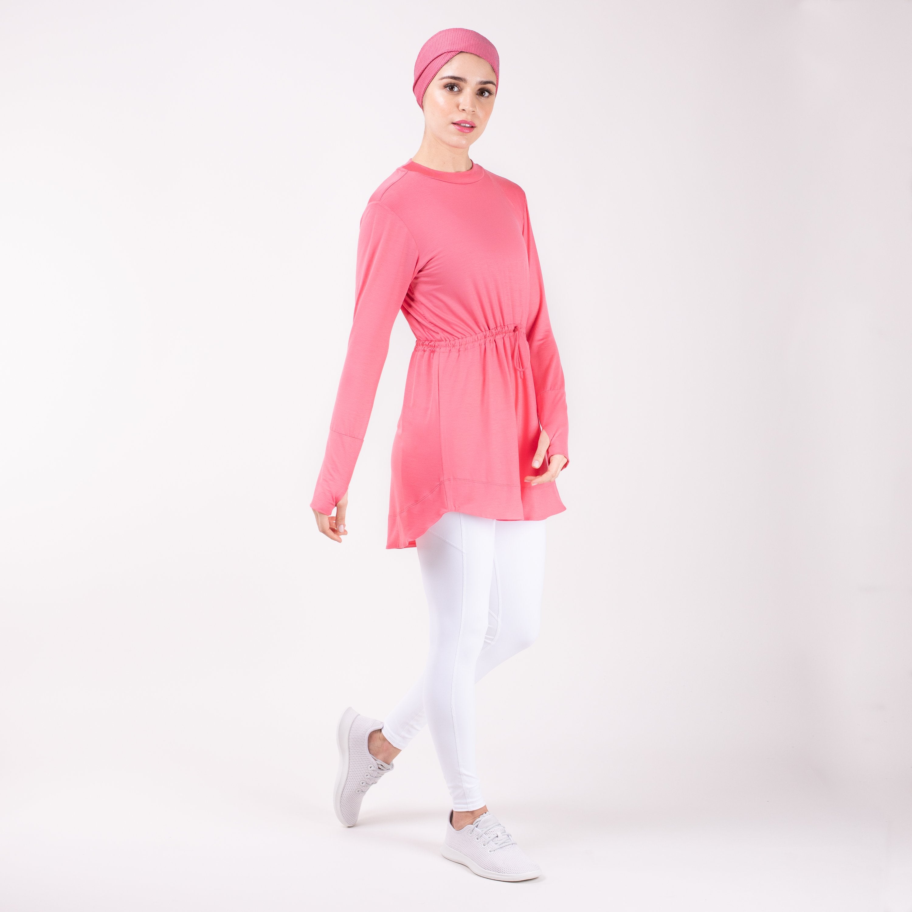 Woman walking to the right wearing the modest, berry pink HAWA drawstring tee shirt and white leggings in front of a white back drop.