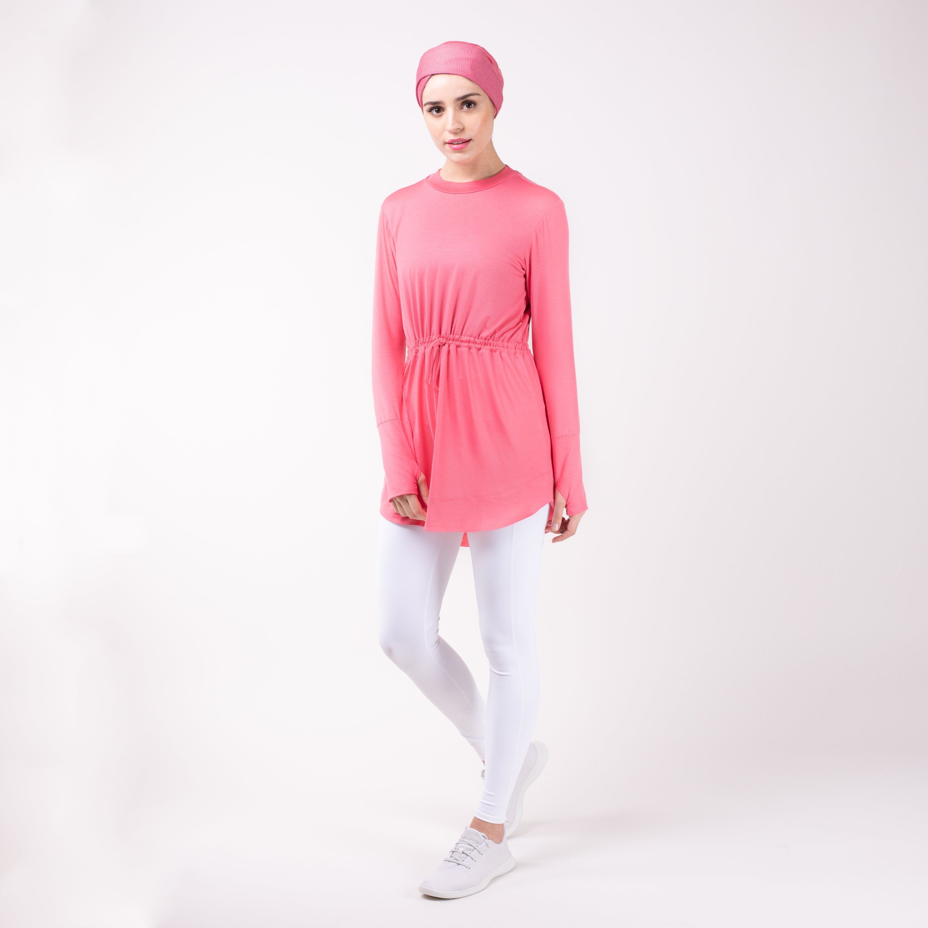 Woman standing with one leg bent wearing the modest, berry pink HAWA drawstring tee shirt and white leggings in front of a white back drop.