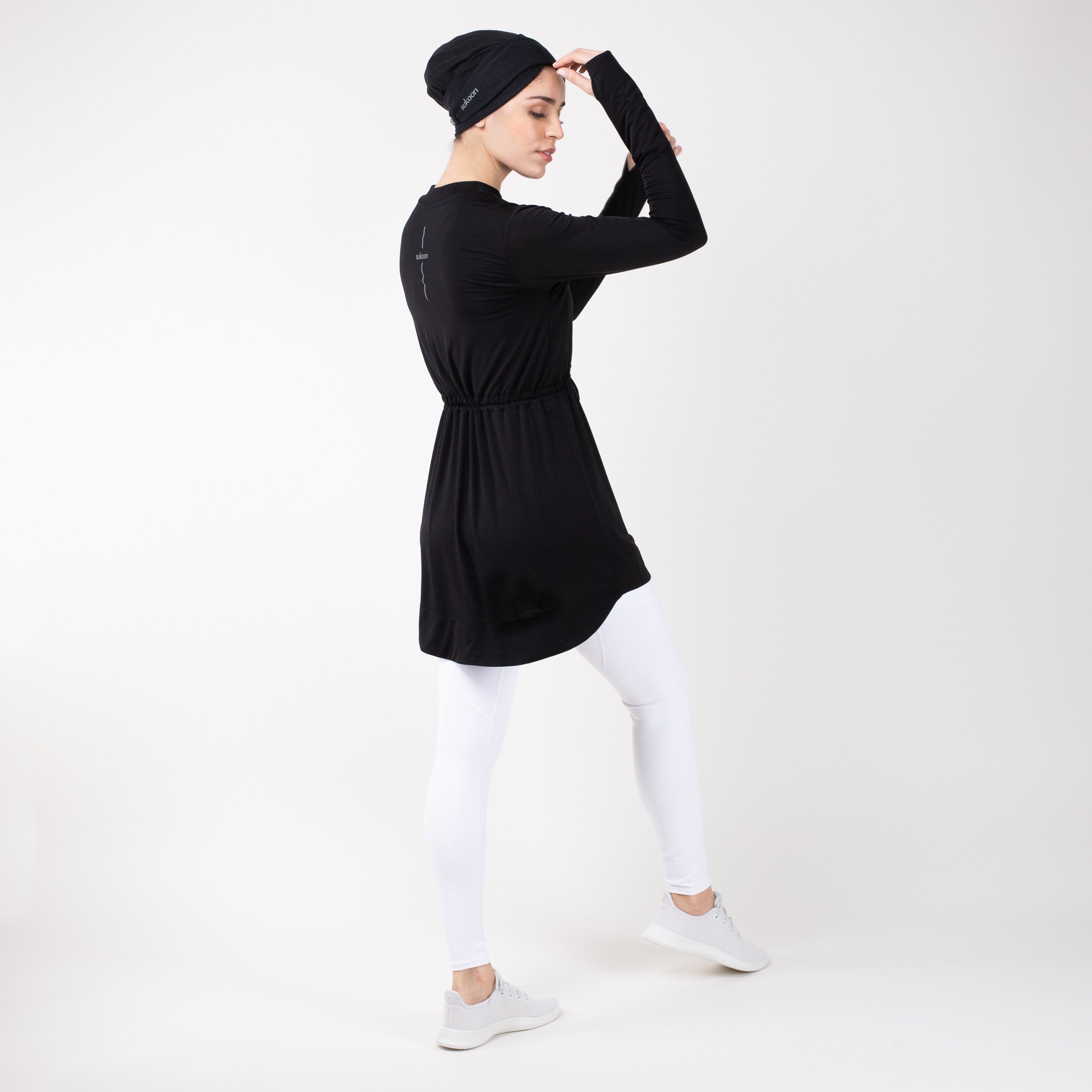 Back of woman in modest, black HAWA drawstring tee shirt and white leggings in front of a white backdrop.