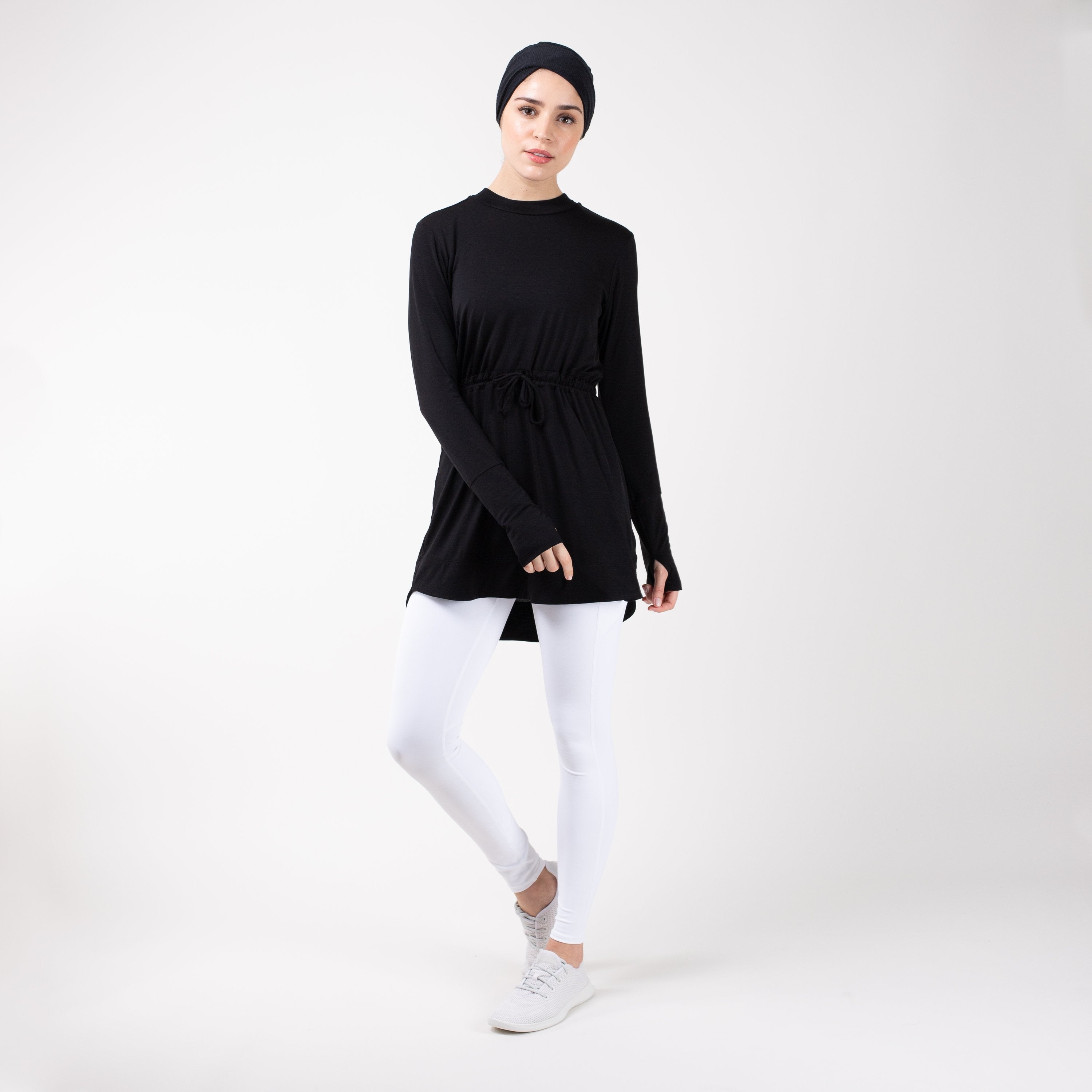 Woman in modest, black HAWA drawstring tee shirt and white leggings in front of a white backdrop.