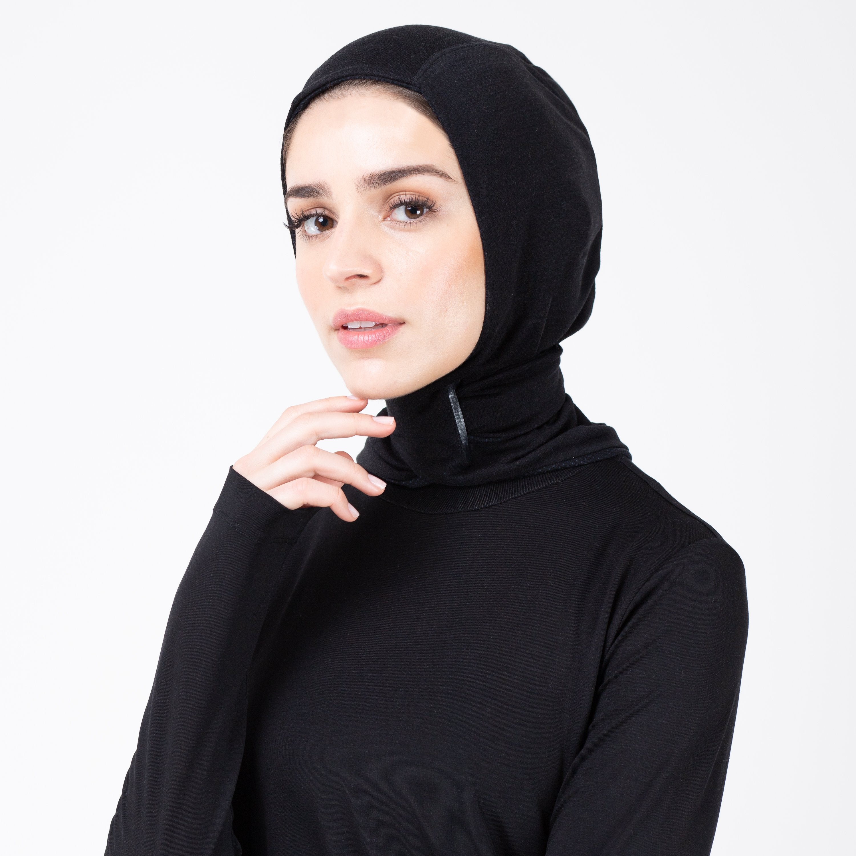 Woman facing left in black shirt and matching black HAWA hijab touching her face.