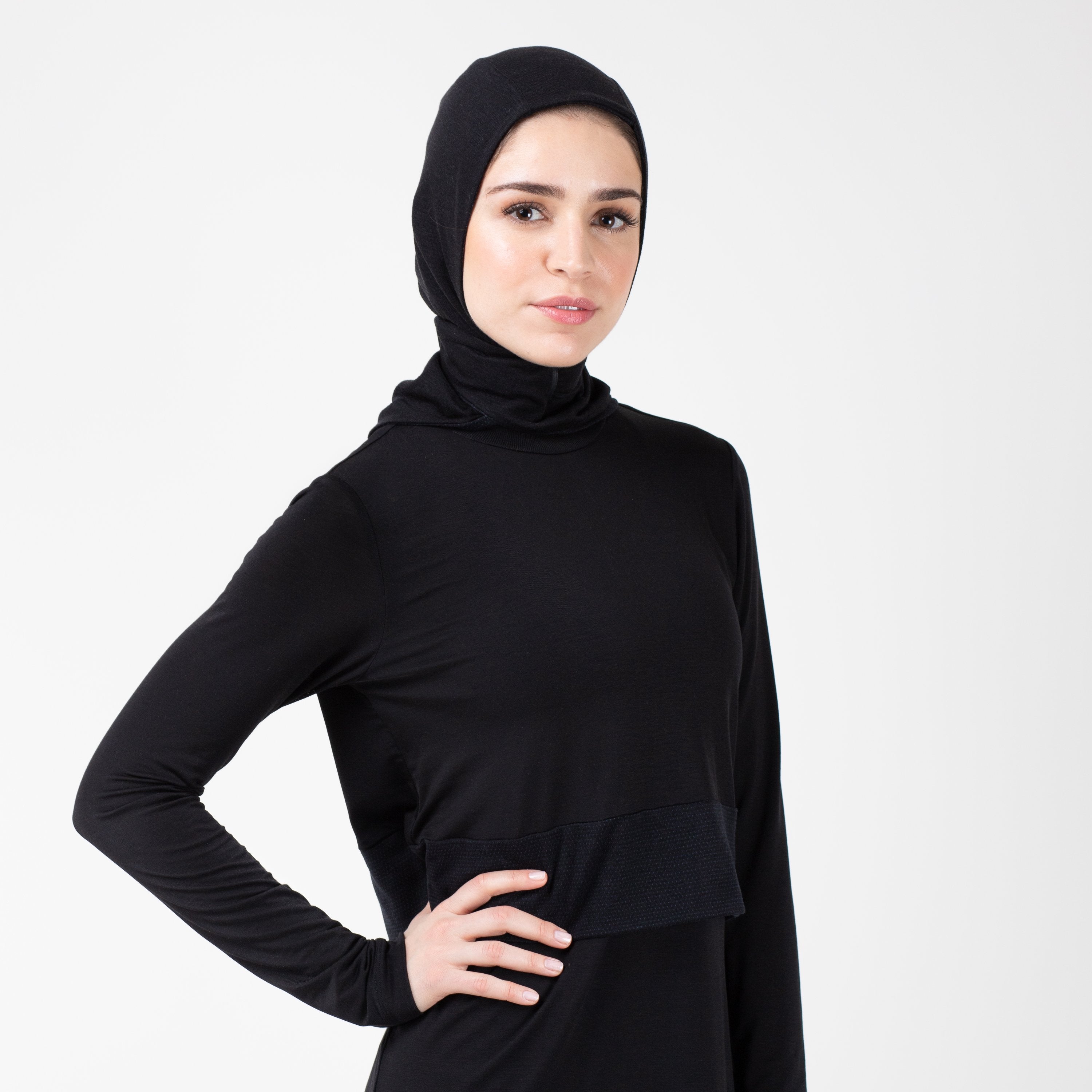 Woman facing right in a black shirt with a matching black HAWA hijab with her right hand on her hip.