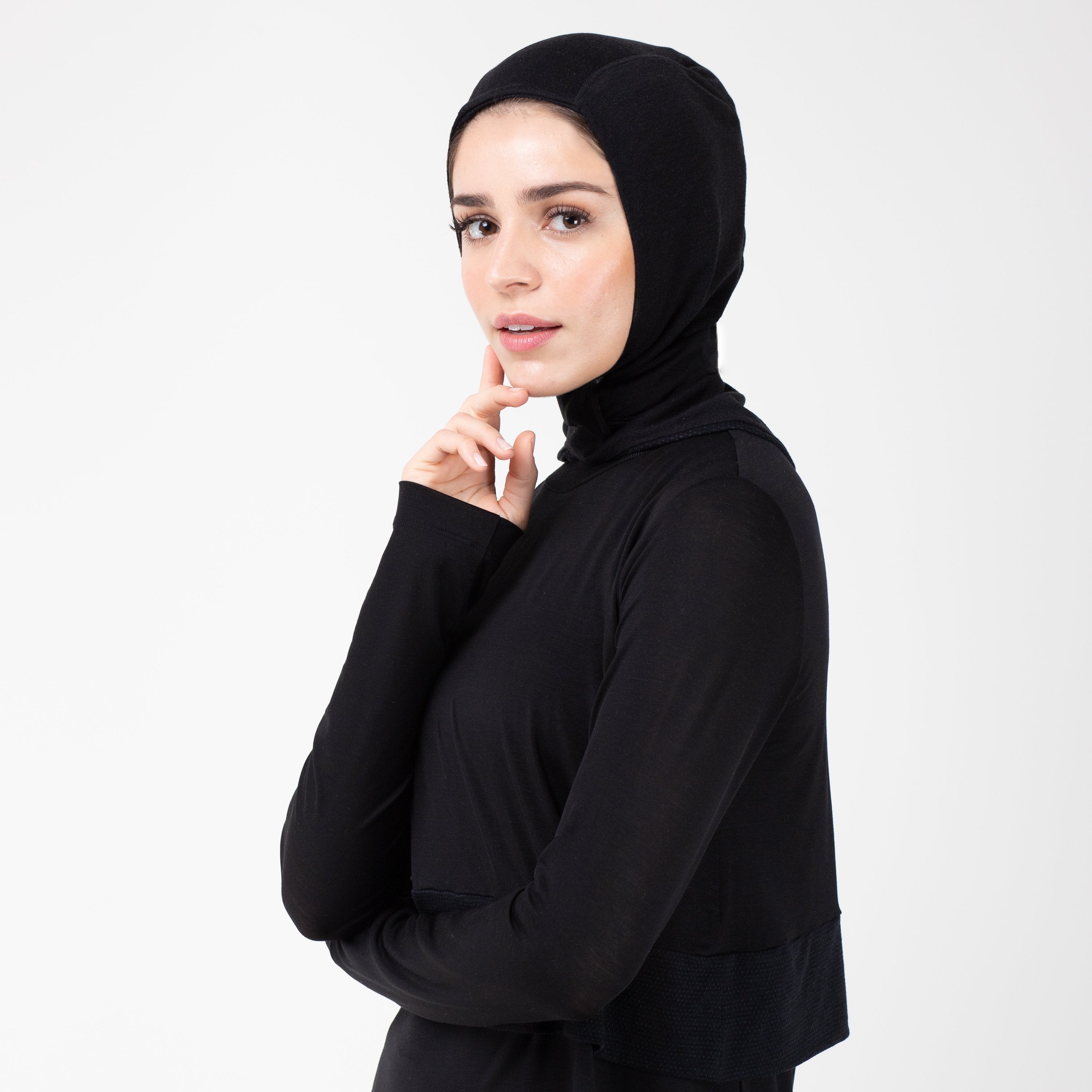 Woman turned to the left wearing a black shirt with a matching black HAWA hijab touching her face with her right hand.