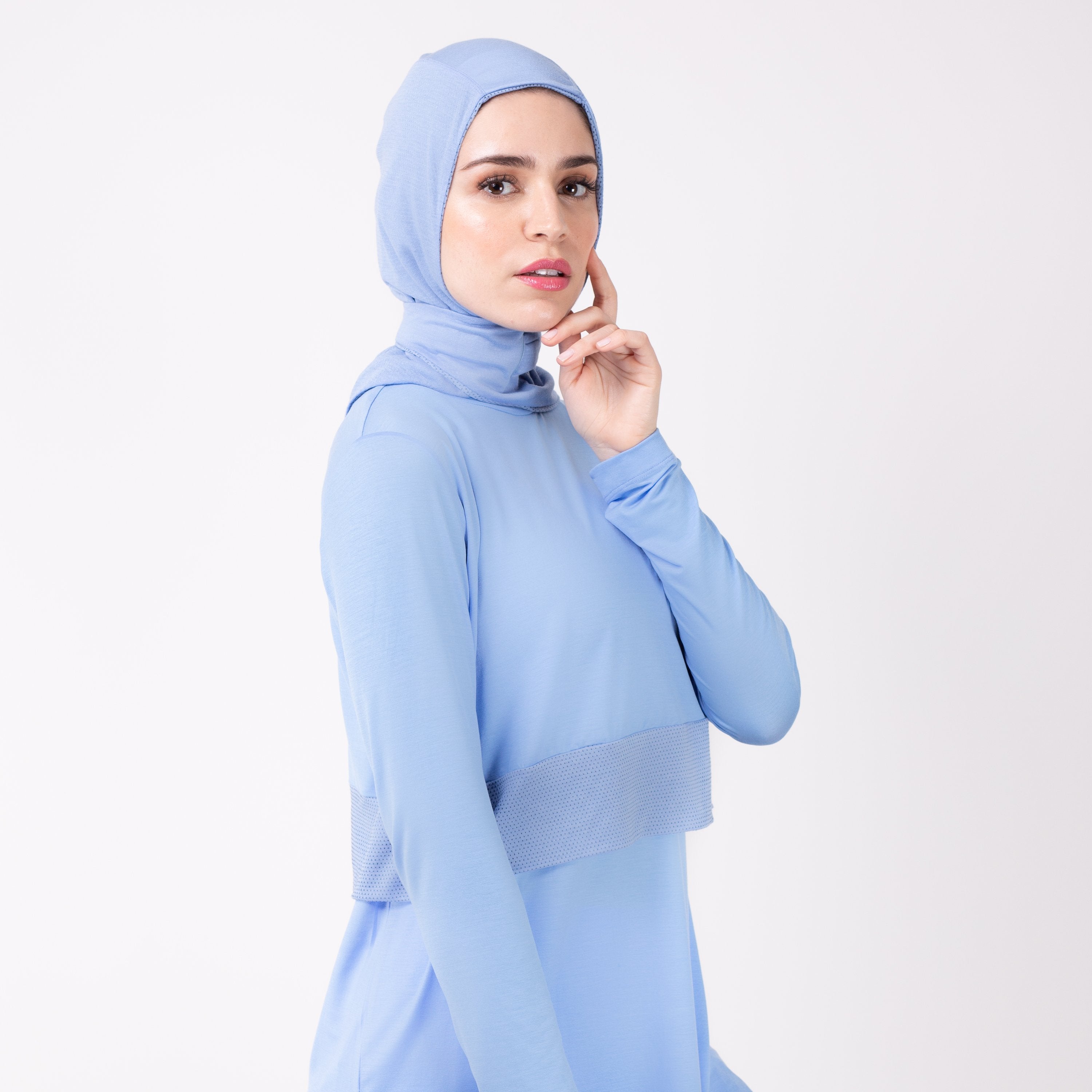 Woman facing right in a sky blue shirt with a matching sky blue HAWA hijab, touching her face with her left hand.