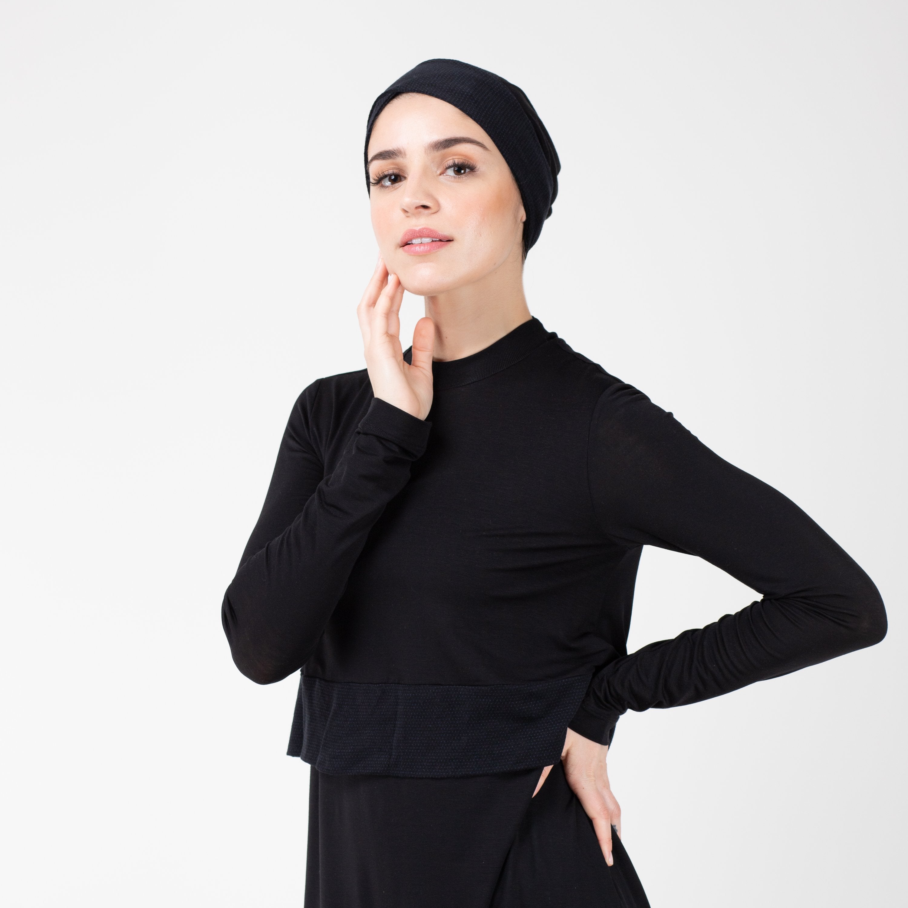 Woman facing left in black shirt with matching black HAWA headwrap, with her right hand touching her face and her left hand on her hip.